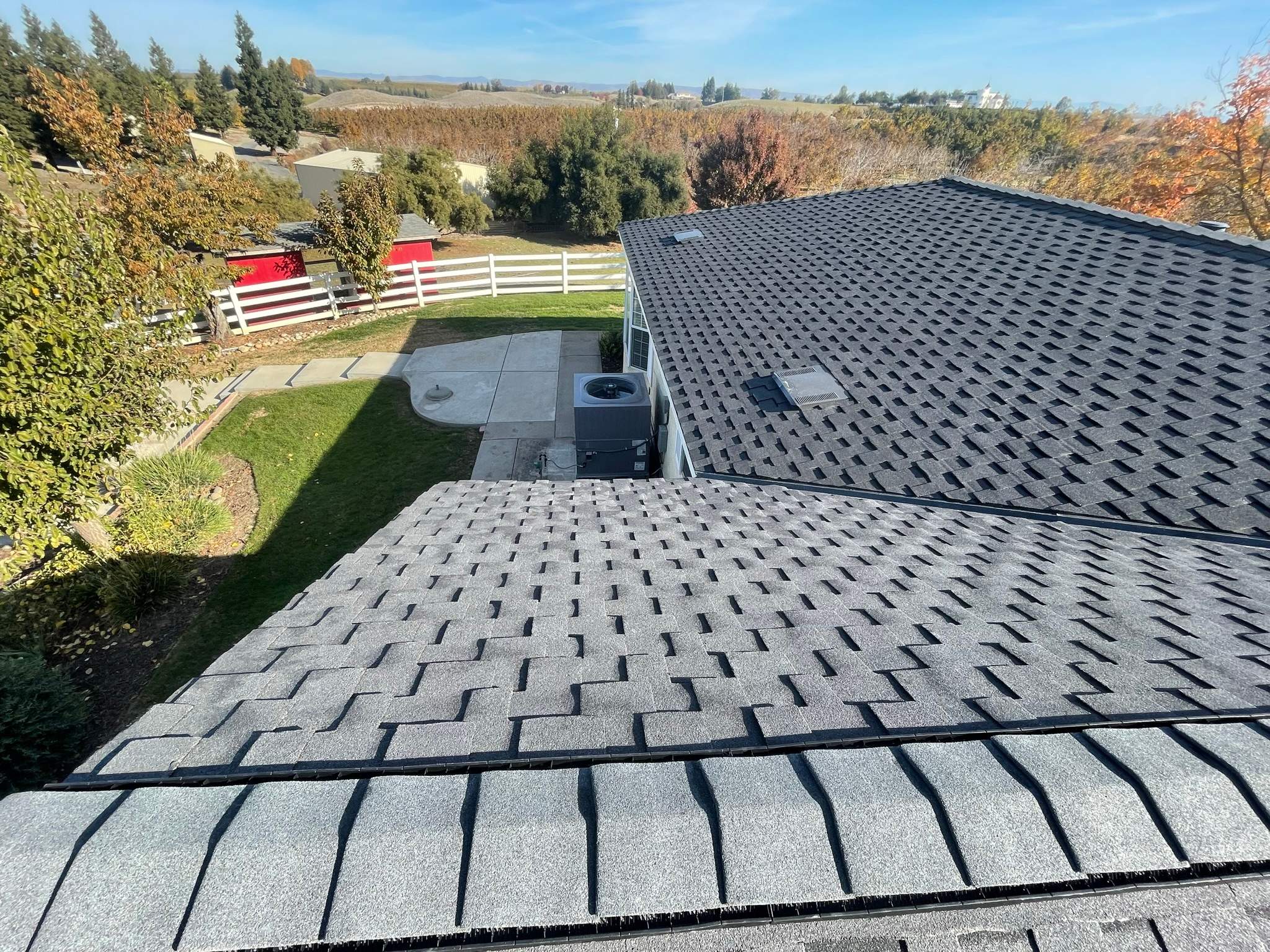 View of two gray shingled roofs with an outdoor HVAC unit between them and a fenced yard with trees and a red shed in the background.