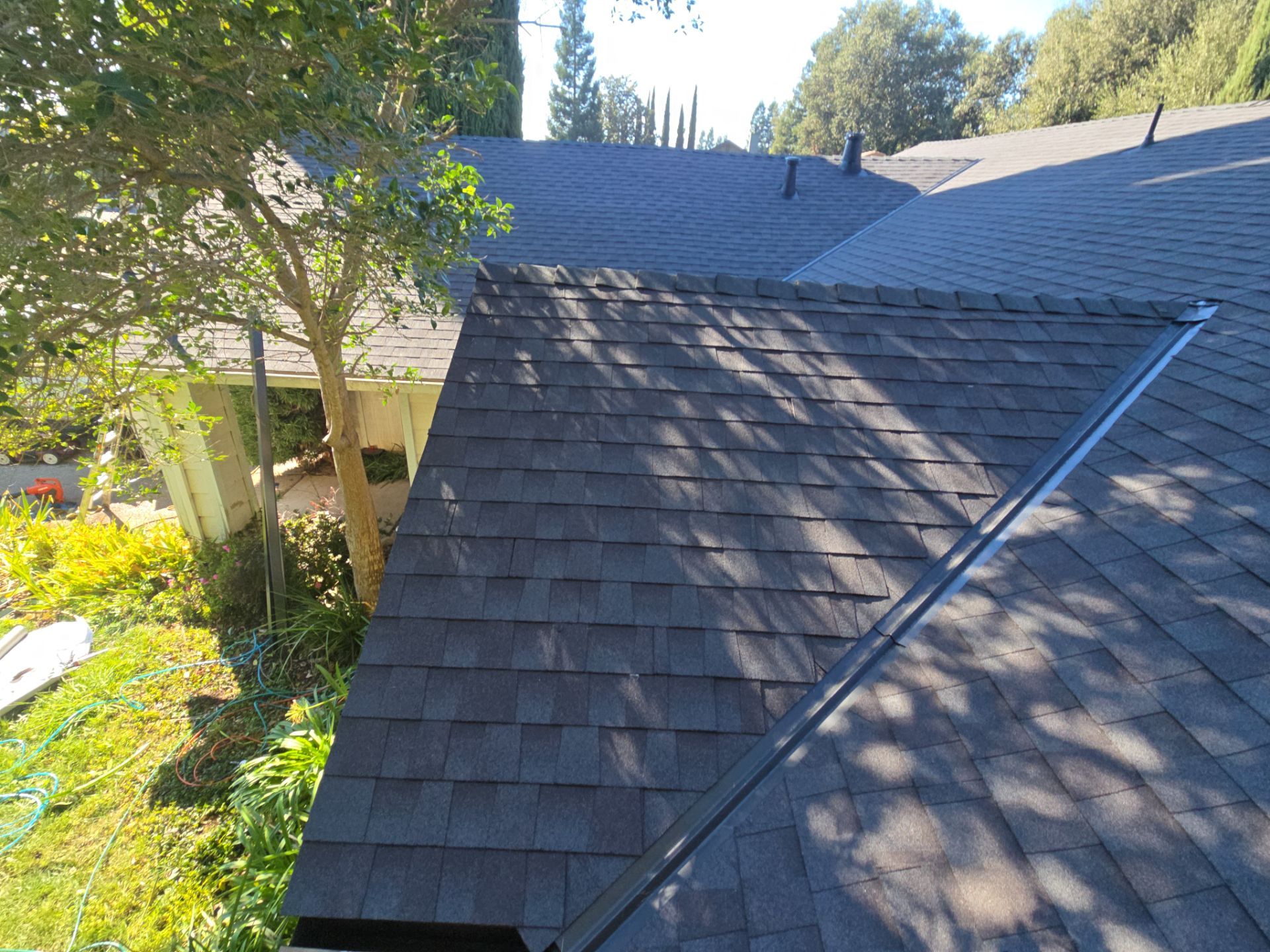 Close-up view of a dark gray shingled roof section with tree shadows and surrounding greenery.