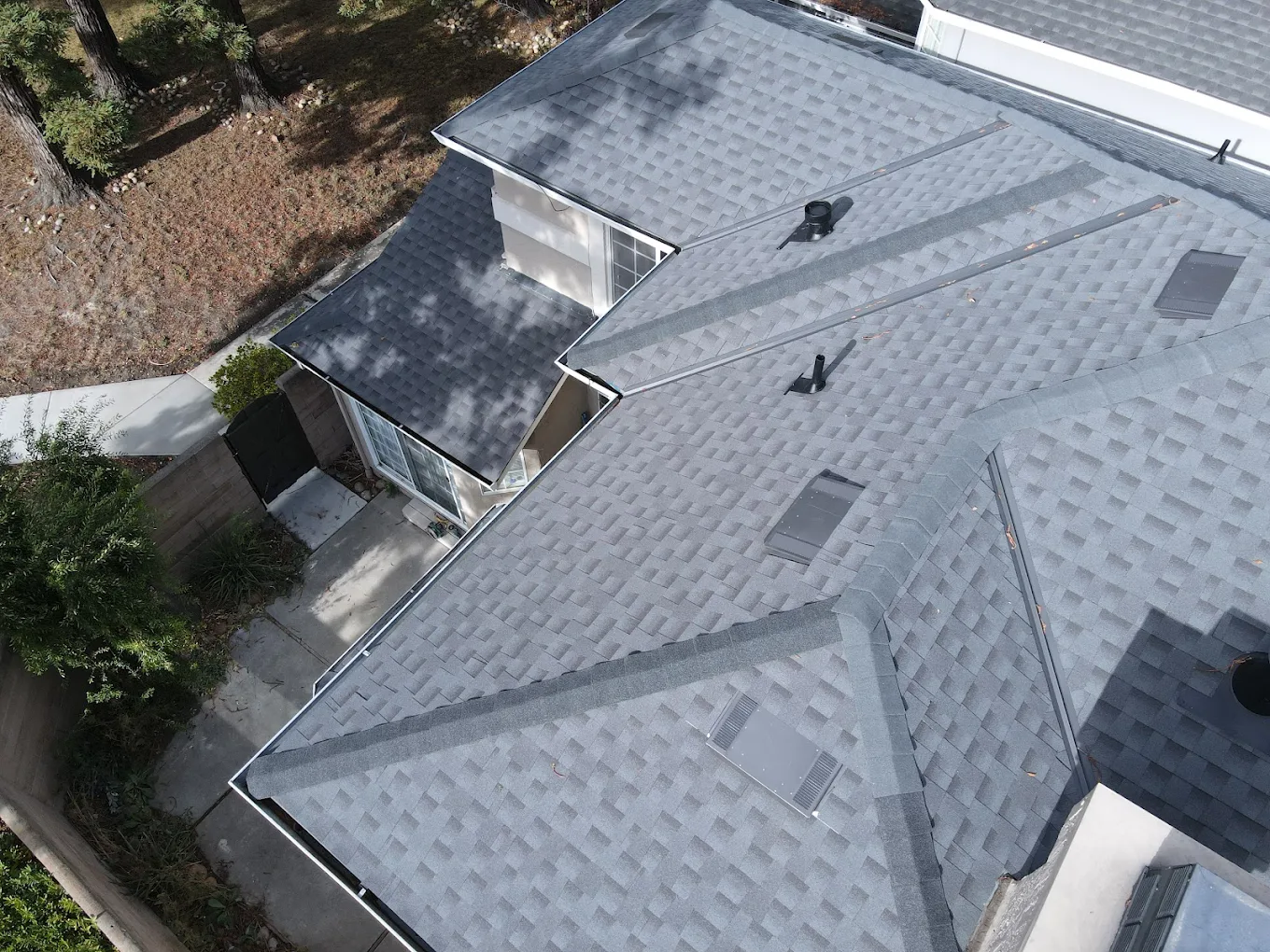 Aerial view of a house roof with gray shingles, vents, and pipes, alongside a small backyard with trees and a sidewalk.