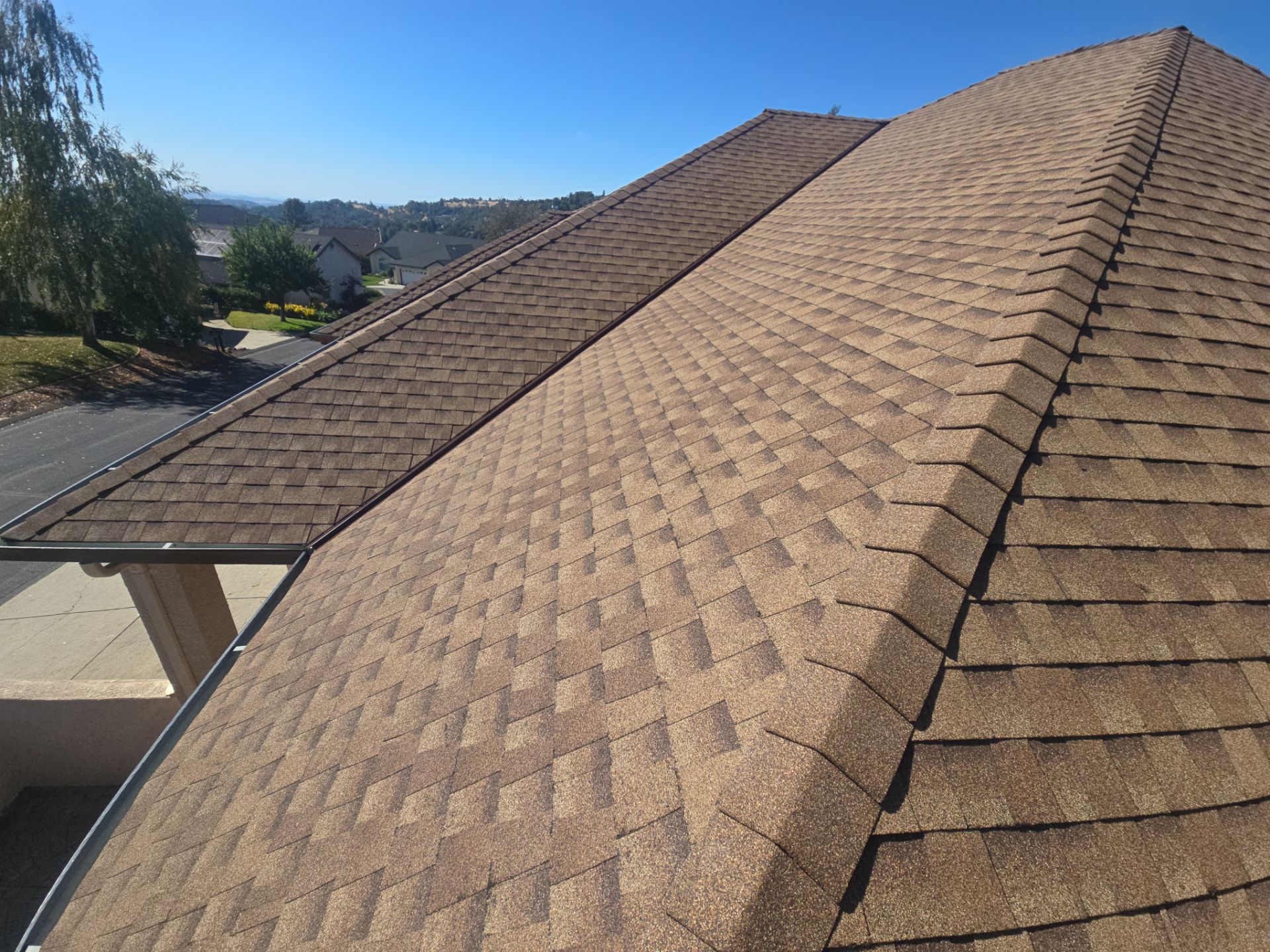 Close-up view of a brown shingled roof with a clear blue sky and suburban houses in the background.