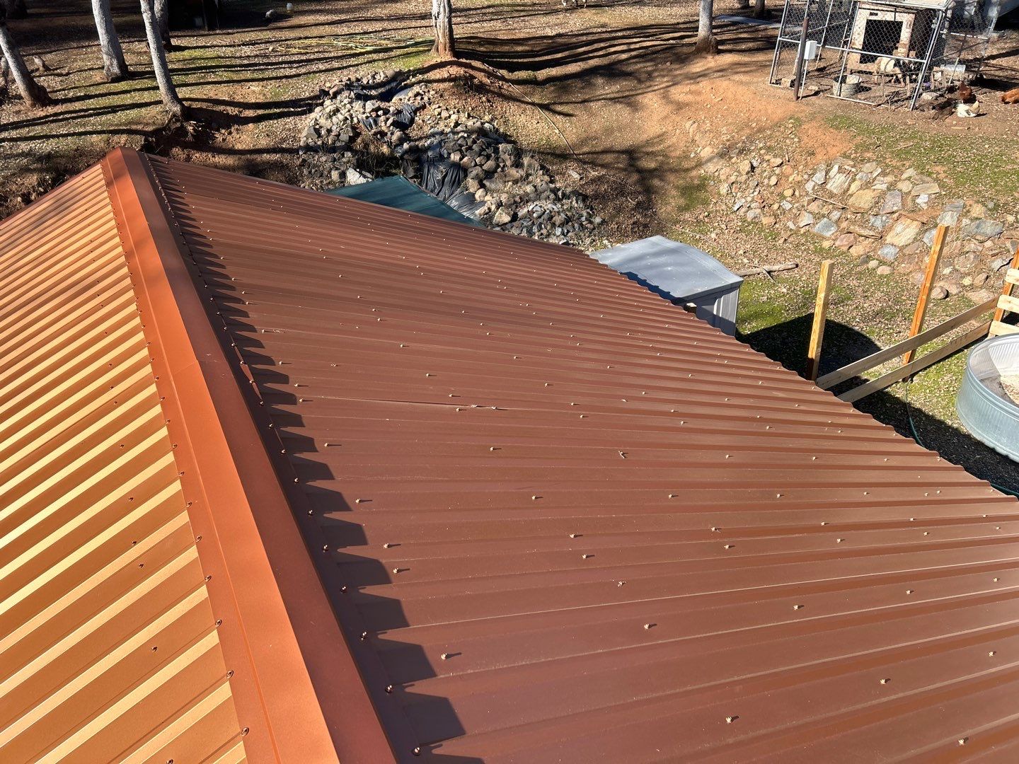 Close-up view of a brown metal roof with screws, seen from above with a backyard and rock wall in the background.
