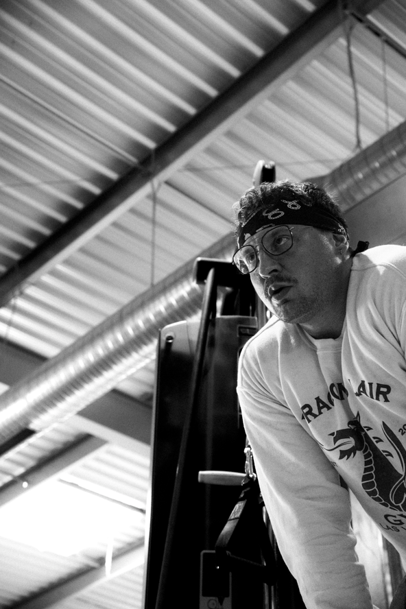 Homme portant un bandana et des lunettes, habillé d'un sweat avec un dragon, dans une salle de sport industrielle.