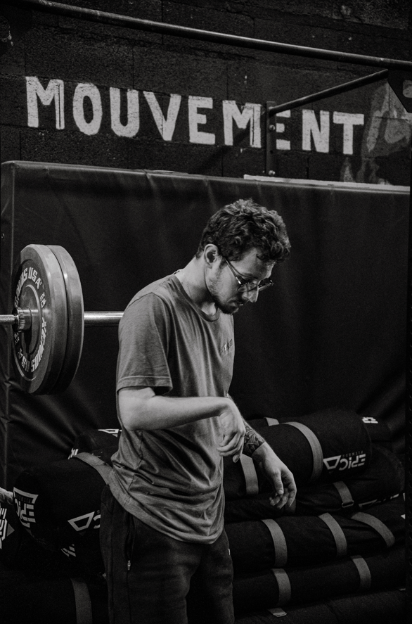 Homme portant des lunettes dans une salle de sport, debout près d'une barre avec des poids, avec le mot "MOUVEMENT" écrit en grand sur le mur derrière lui.