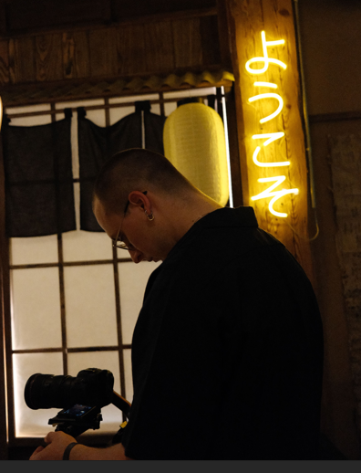 Homme regardant un appareil photo dans un intérieur au décor japonais avec un panneau lumineux affichant ようこそ.