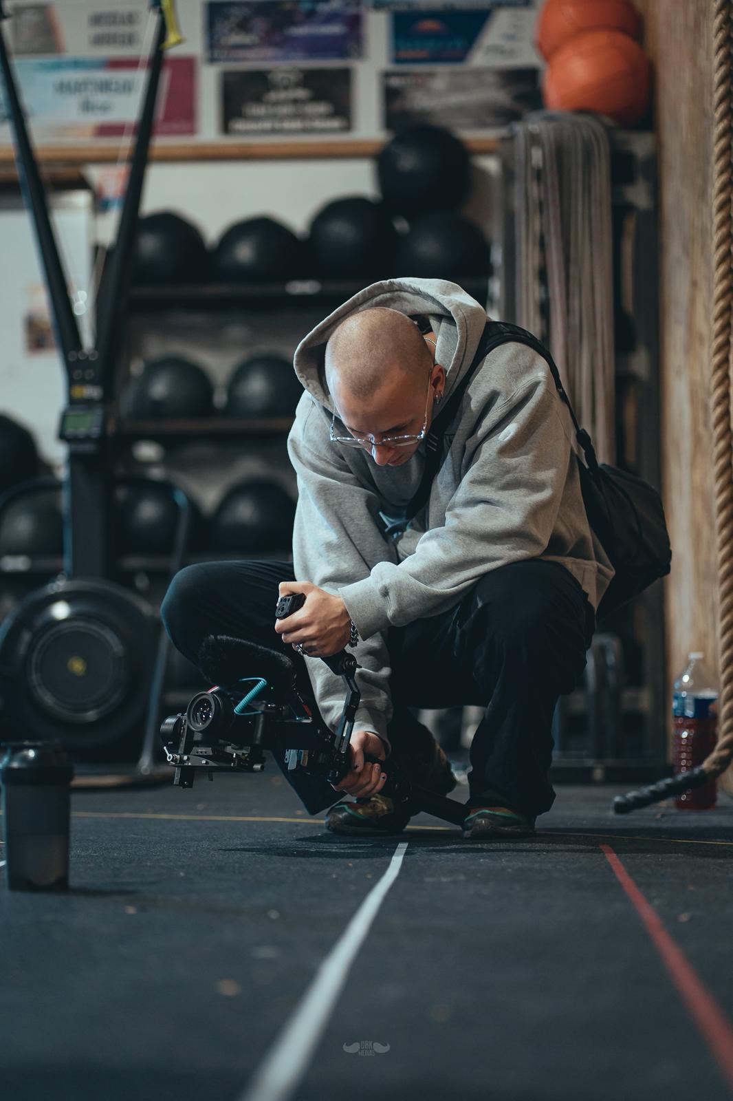 Un homme en sweat à capuche gris accroupi dans une salle de sport, tenant une caméra vidéo professionnelle au sol.