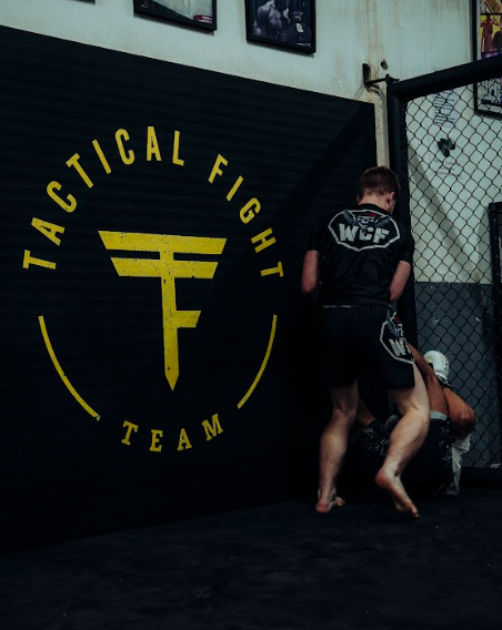 Deux hommes s'entraînent en arts martiaux mixtes dans une cage, à côté d'un mur avec un grand logo jaune 'Tactical Fight Team'.
