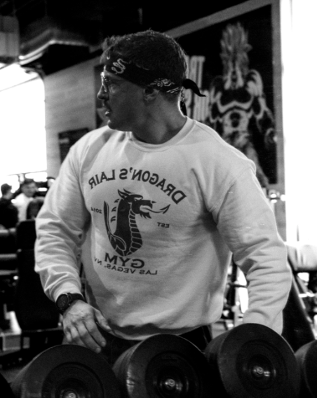 Homme portant un bandeau et un sweat Dragon's Lair Gym soulevant des haltères dans une salle de sport.