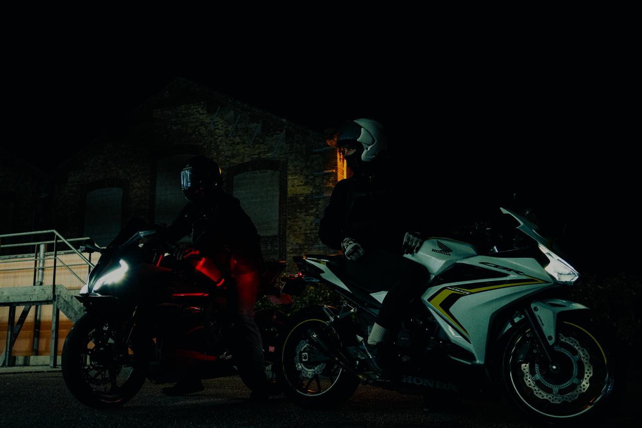 Deux motards portant des casques sur des motos sportives, l'une blanche avec des détails jaunes et l'autre sombre avec les phares allumés, la nuit devant un bâtiment en briques.