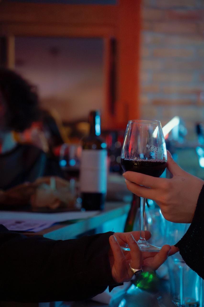 Main tenant un verre de vin rouge offert par une autre personne dans un cadre intérieur tamisé à l'ambiance chaleureuse.