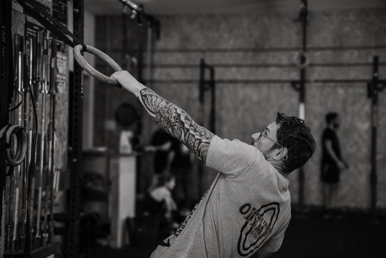 Un homme tatoué portant des lunettes s'entraîne avec des anneaux de gymnastique dans une salle de sport.