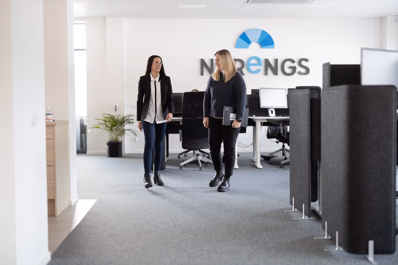 Två professionella kvinnor i affärskläder står på modernt kontor med Norengs logotyp på väggen - Personlig ekonomisk rådgivning Gävle