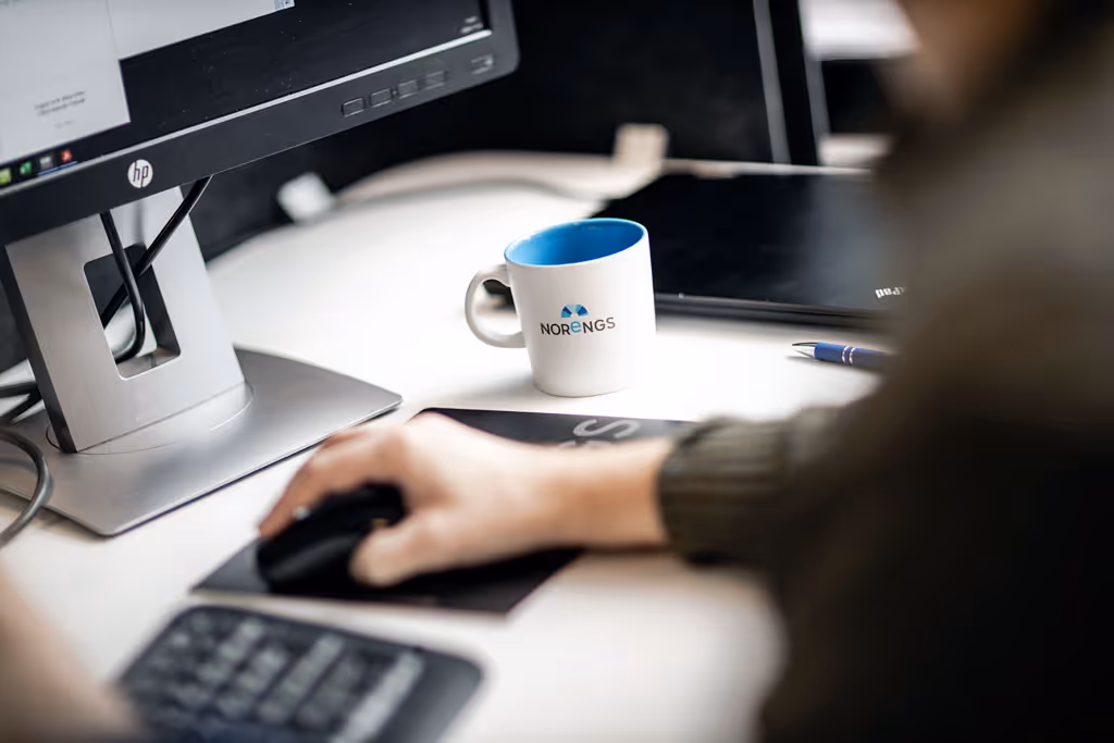 Person arbetar vid dator med vit Norengs kaffekopp på skrivbord, hand på datormus, HP-skärm - Modern redovisning och ekonomisk rådgivning Gävle
