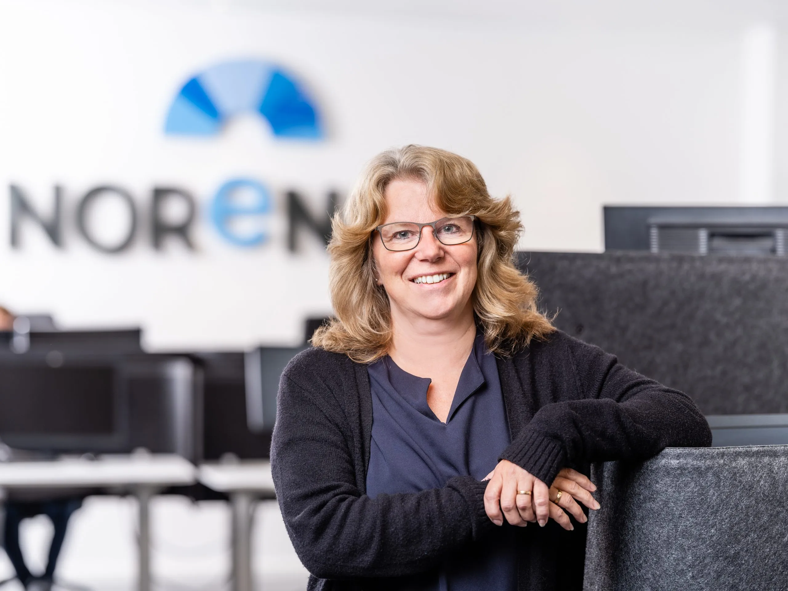 Leende kvinna med glasögon och ljuslockigt hår lutar sig mot en grå skärm på ett redovisningsbyrå kontor i sälen.