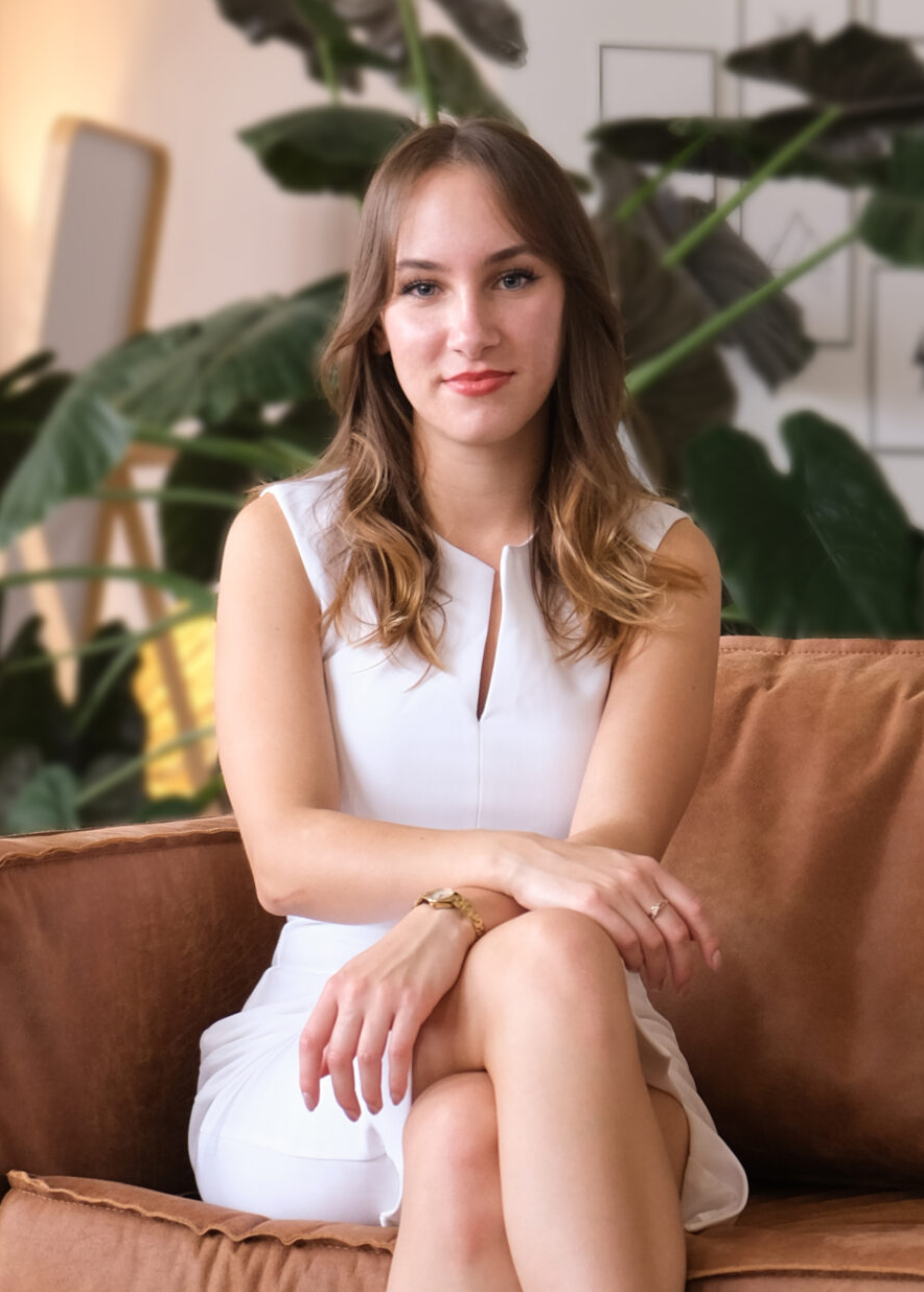 Annabell Scheibel, Gründerin der Askara Marketing GmbH, sitzt in einem weißen Kleid und roten High Heels auf einem braunen Ledersofa in einem modern eingerichteten Raum mit Pflanzen im Hintergrund.