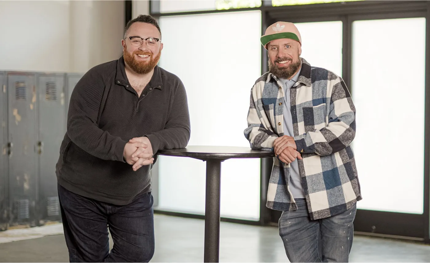 Two smiling men standing at a high table in a modern office space with large windows in the background