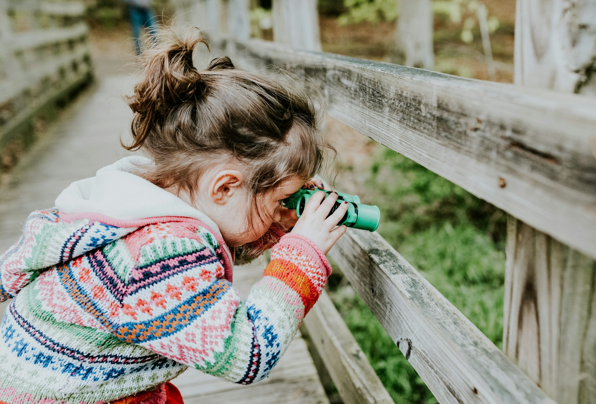 A curly-haired girl looks through toy binoculars by a wooden fence