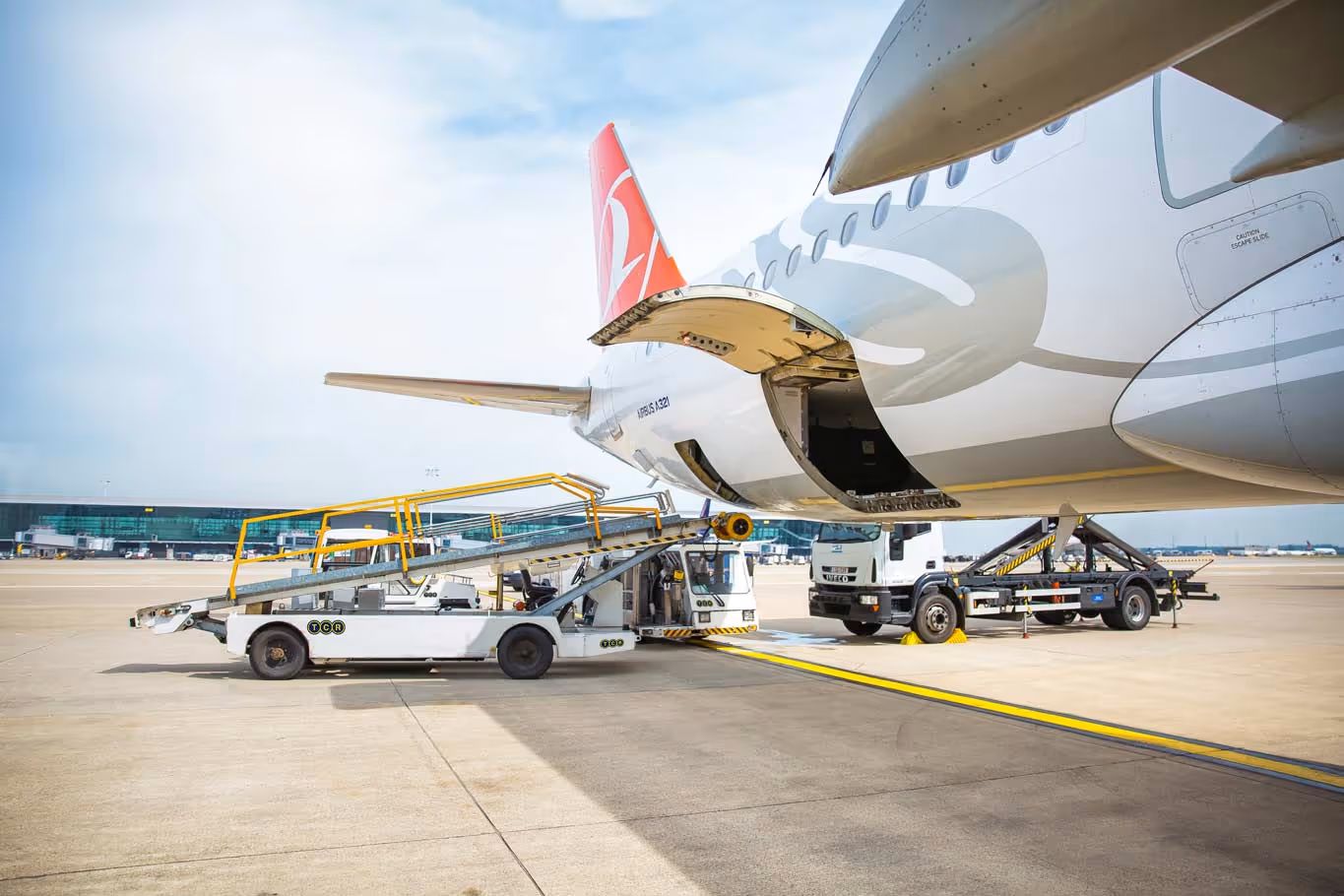 Airport ground support equipment loading cargo into the open rear cargo door of a parked commercial airplane with a red tail fin under a partly cloudy sky.