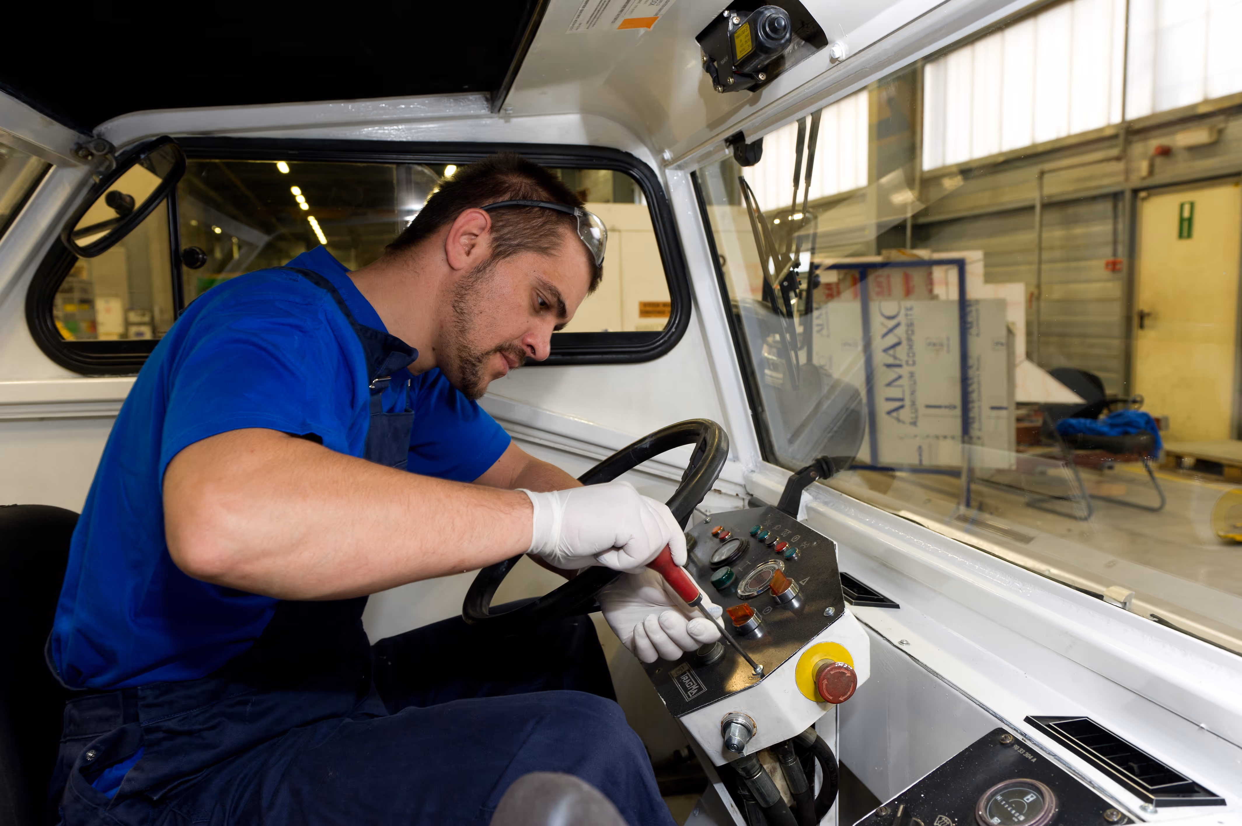 Technician wearing gloves and safety glasses using a screwdriver to repair the control panel inside a vehicle cab.