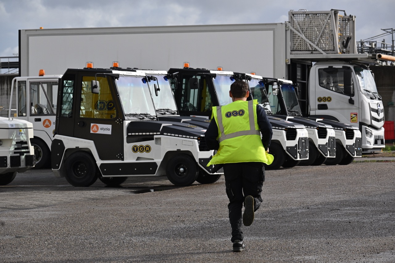 Man in a yellow safety vest running towards a line of small utility vehicles and a large truck in an industrial area.