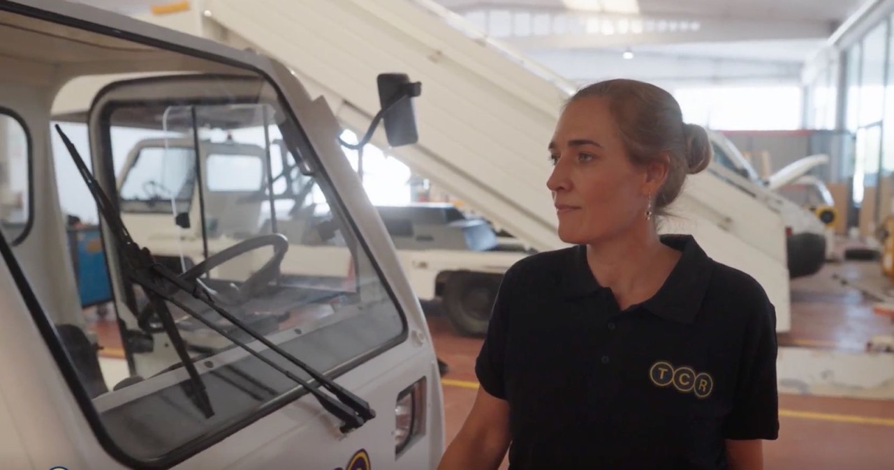 Woman in a black TCR polo shirt standing next to a white utility vehicle inside a workshop.