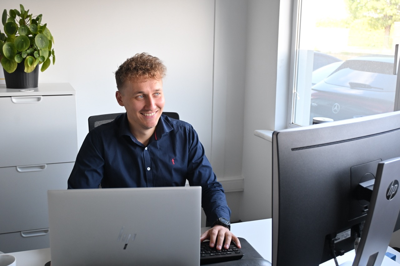 Smiling man with curly hair working at a desk with a laptop and monitor in a bright office.