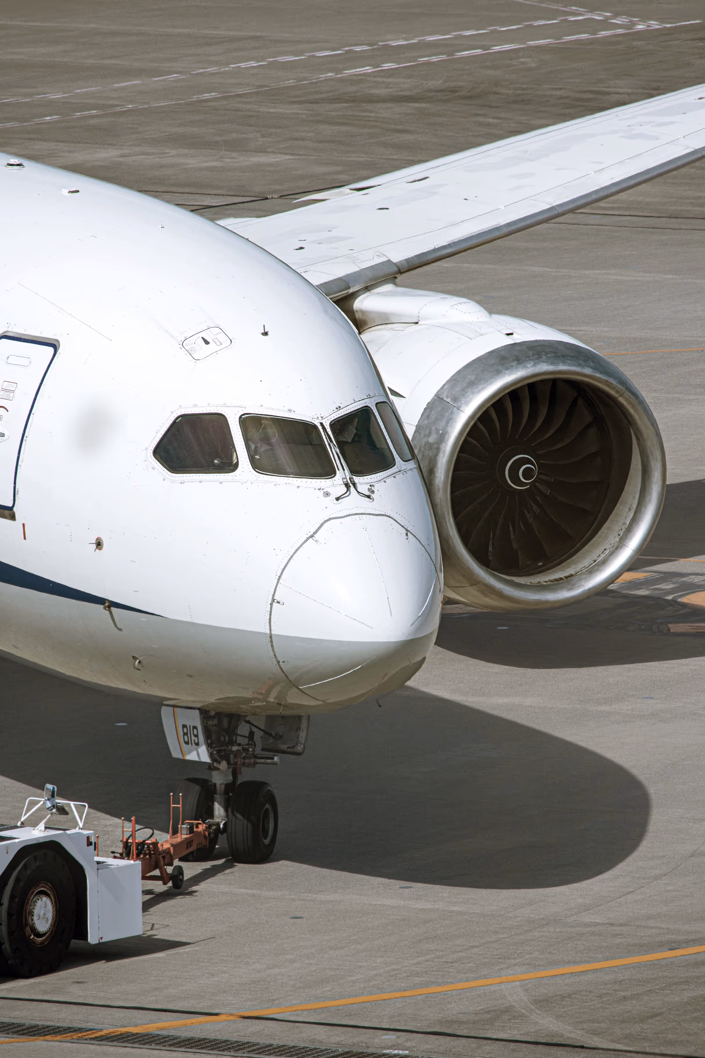Close-up of the nose, cockpit windows, and jet engine of a white airplane on the tarmac with a tug vehicle attached to its front landing gear.