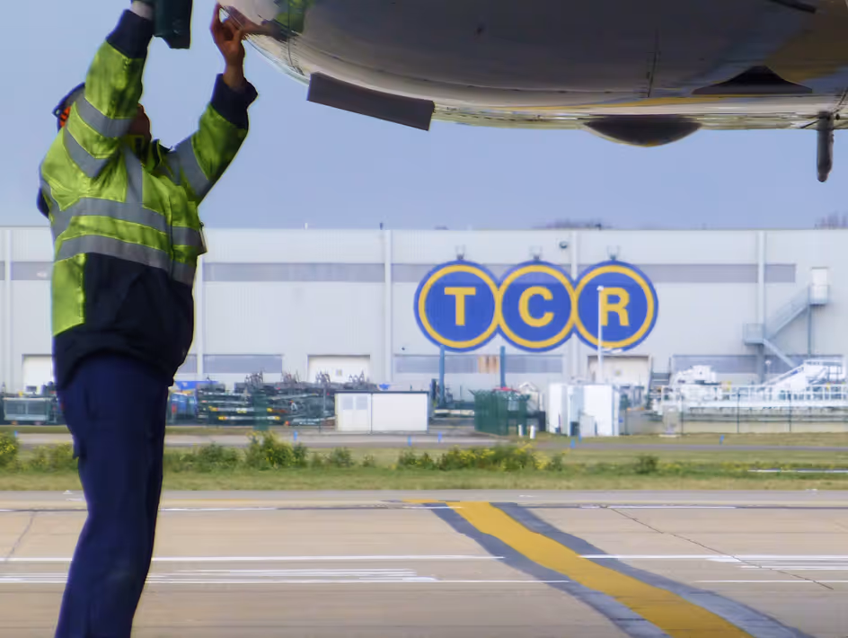 Airport worker in a reflective jacket inspecting the underside of an airplane near large blue and yellow TCR signage.