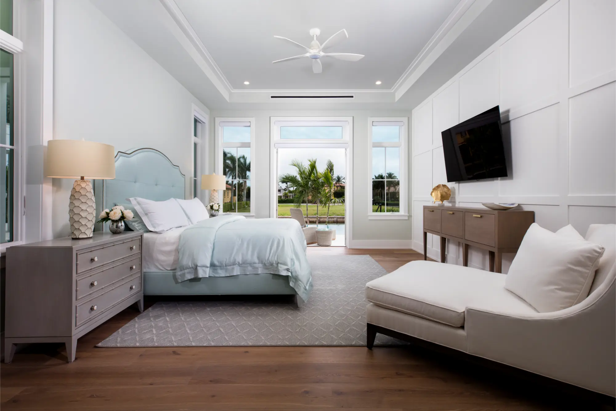 Modern bedroom with a neatly made bed, bedside lamps, a wall-mounted TV, and a chaise lounge.