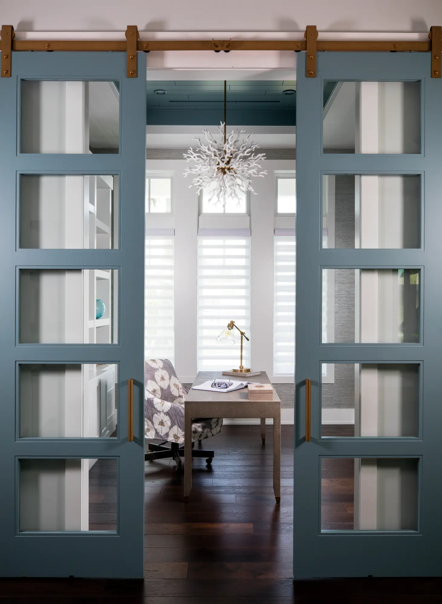 A modern office with glass-paneled sliding doors, featuring a desk, chair, and decorative chandelier.