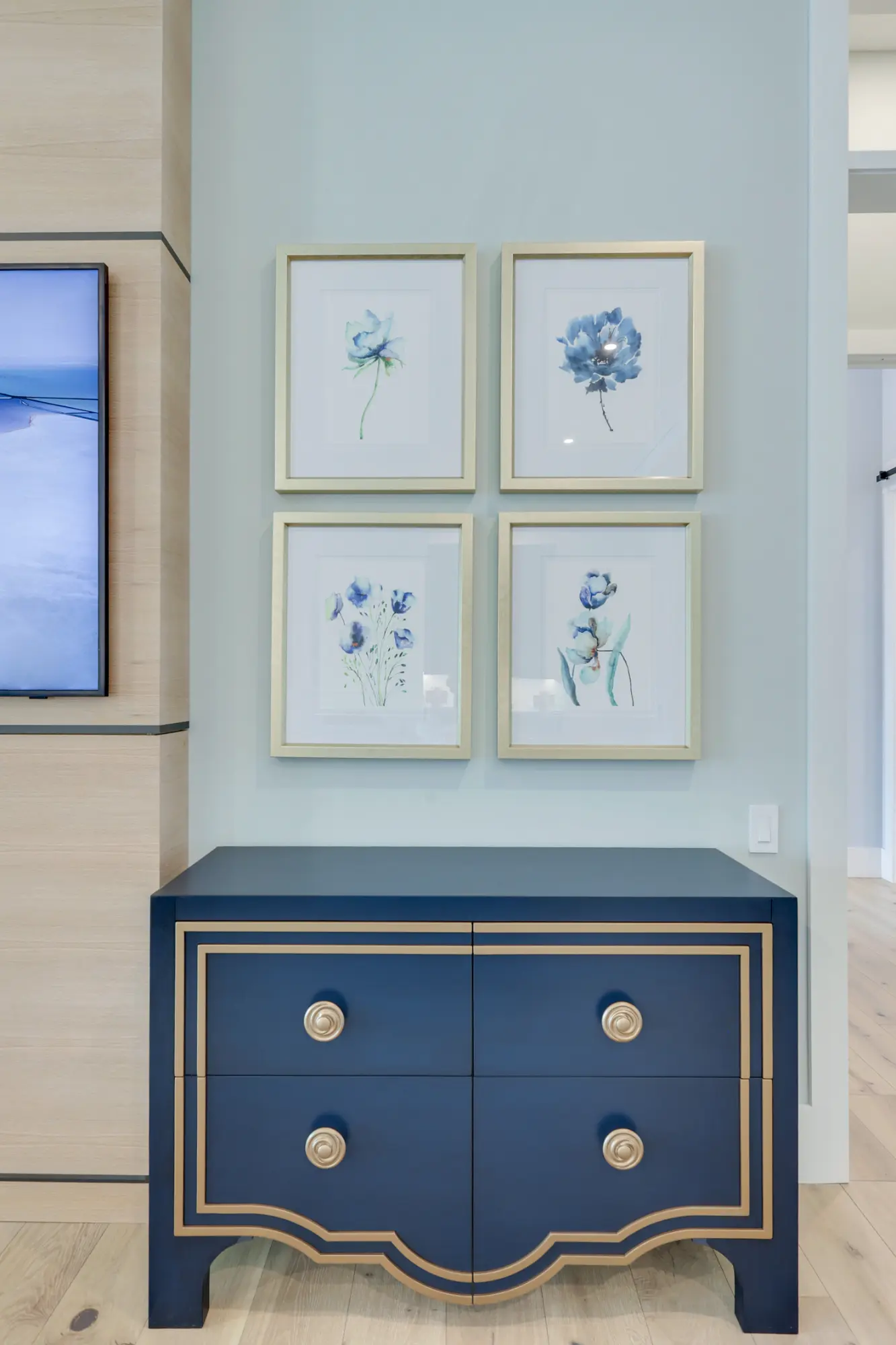 A blue and gold dresser with four floral paintings hanging above it on a light blue wall.