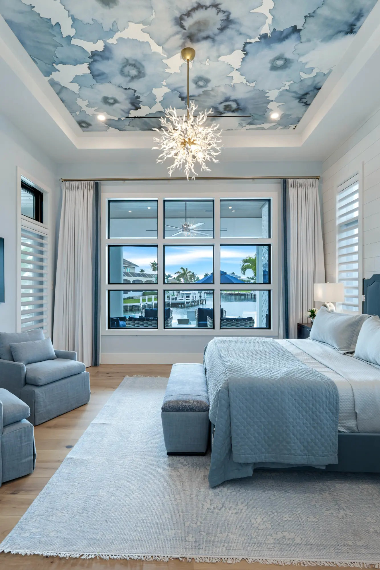 Bedroom with a large bed, gray armchair, and patterned ceiling light fixture. Large windows show outside view.