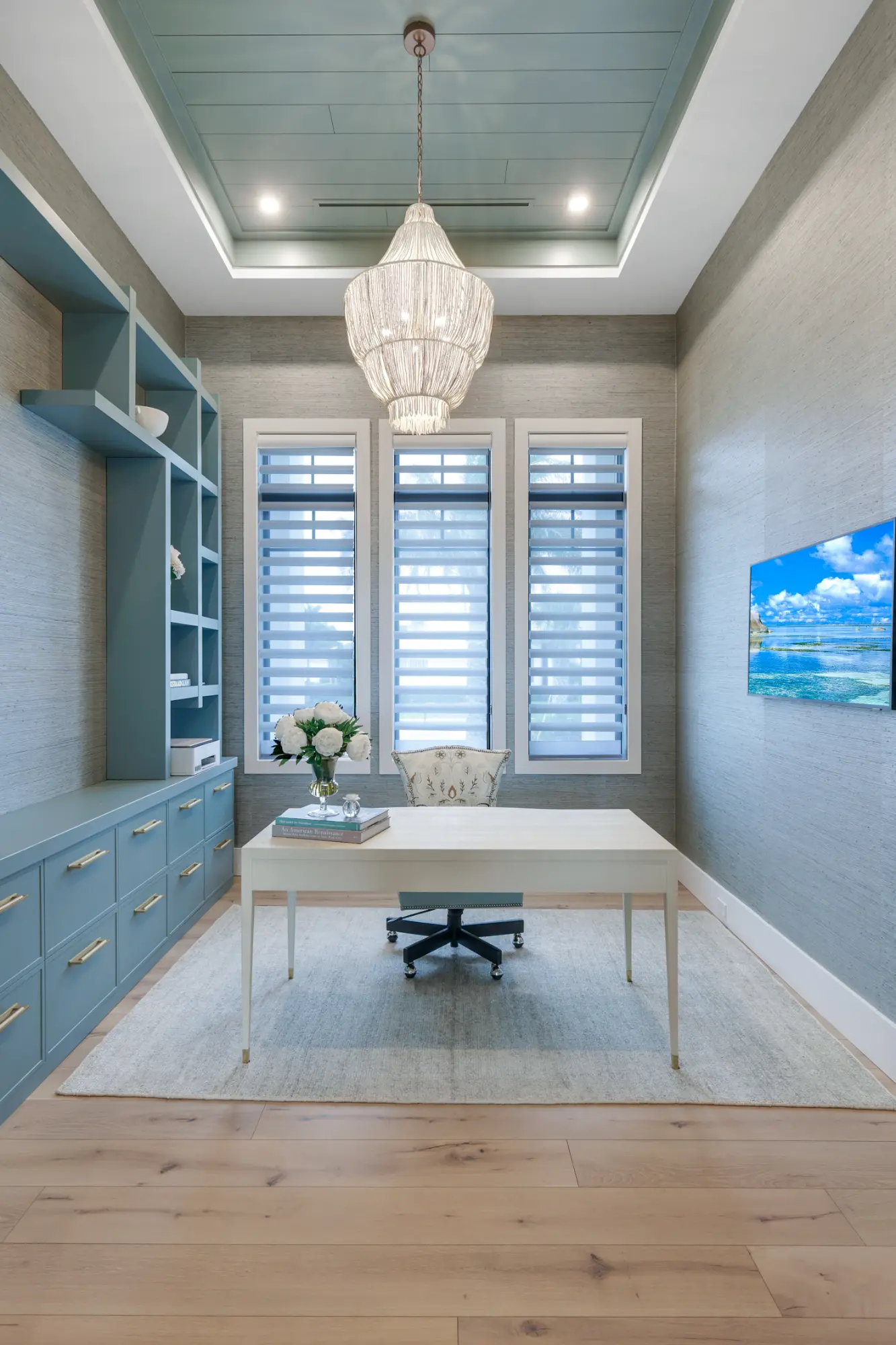 A modern, elegantly decorated home office with a white desk, chair, and a large chandelier above.