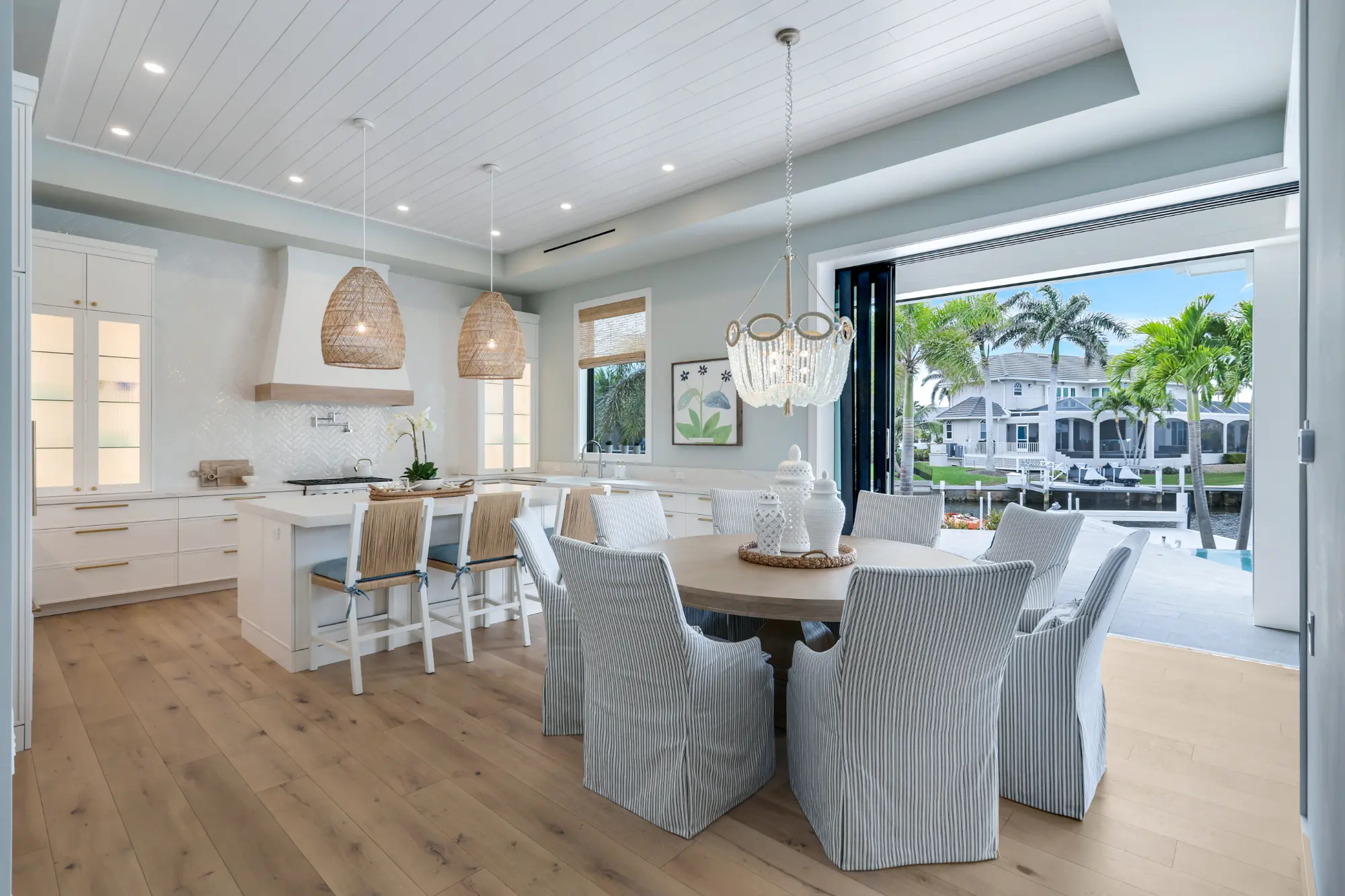 A modern kitchen and dining area with white furniture and large windows overlooking a backyard with a pool.