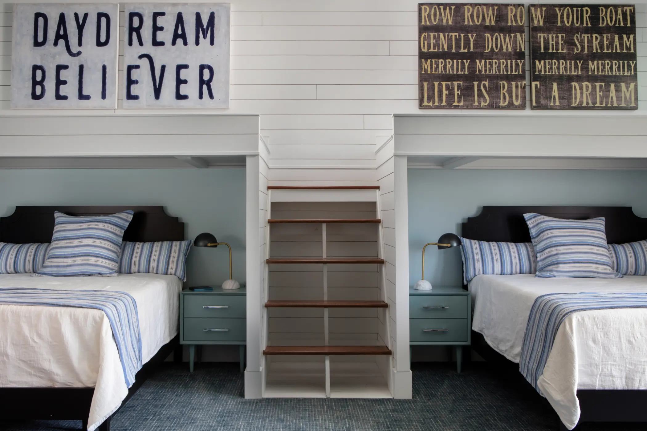 Two beds with striped bedding flank a wooden staircase under decorative wall signs in a cozy bedroom.