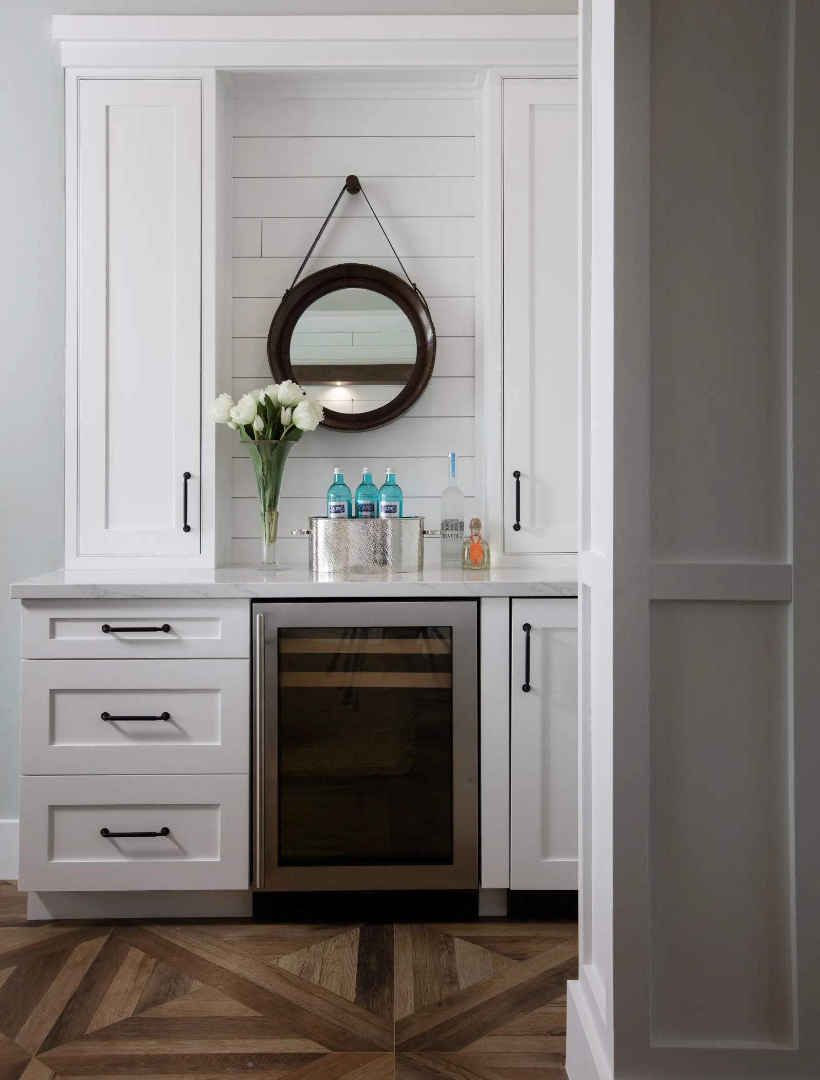 White cabinet with a round mirror, a mini fridge, and a vase of white flowers.