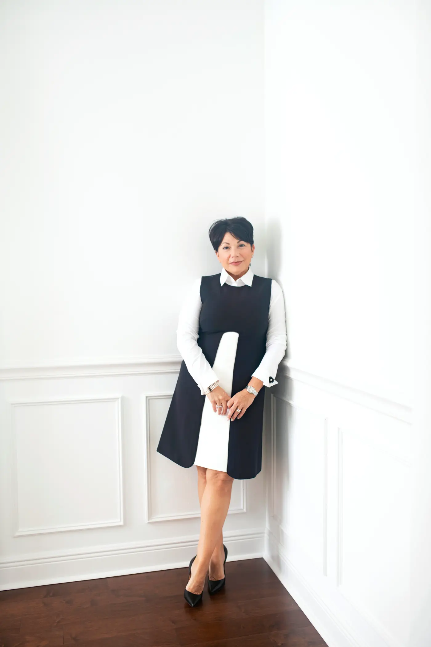 A woman stands against a white wall, wearing a black and white dress, looking forward.