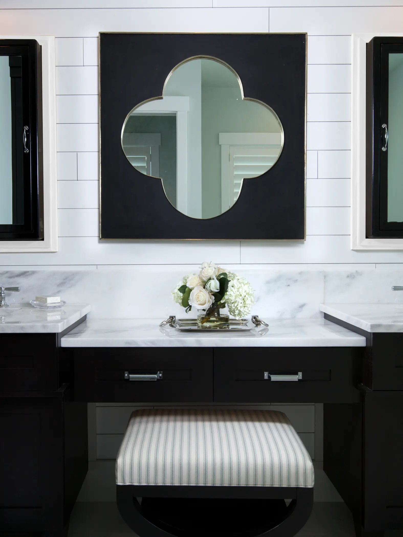 Bathroom vanity with striped cushioned stool, large clover-shaped mirror, and a bouquet of white flowers.
