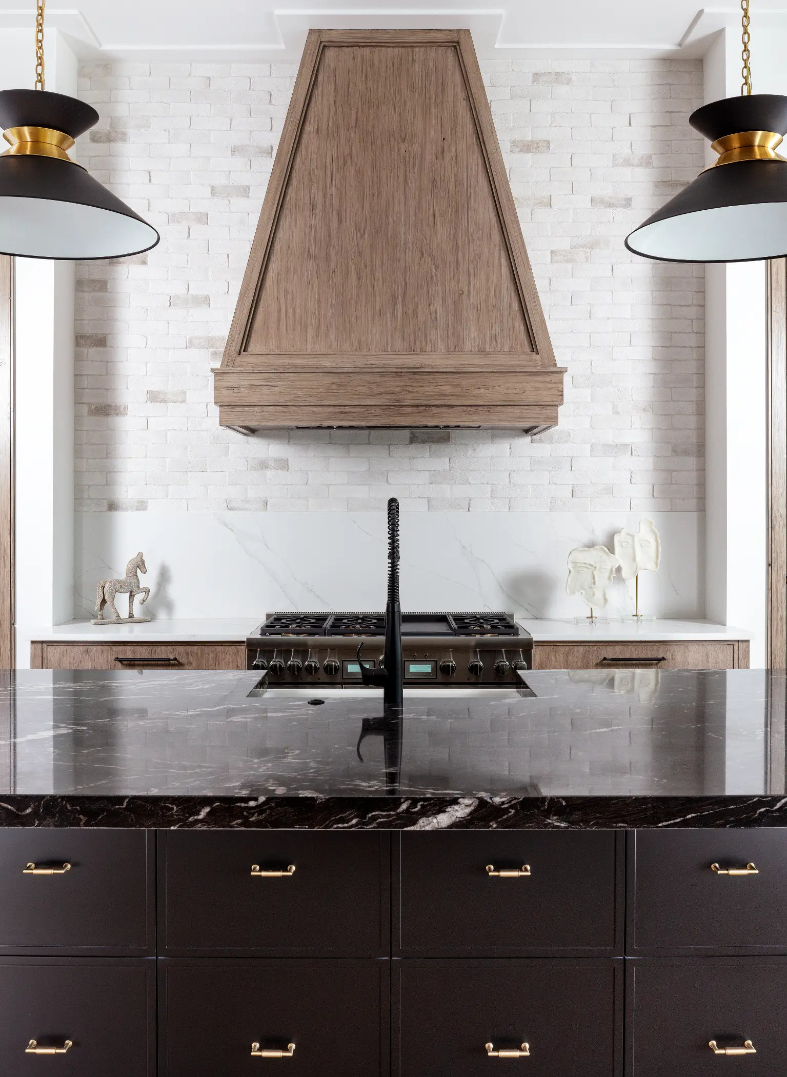 Modern kitchen with dark marble island, wooden hood, and black stove against white brick wall.