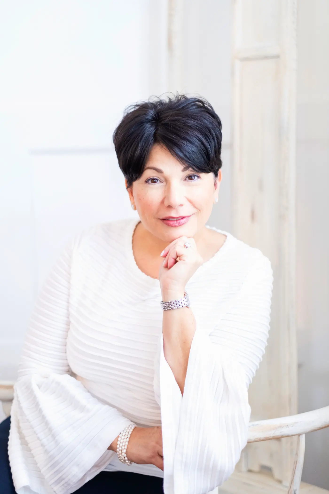 A person with short dark hair sits, wearing a white blouse, resting their chin on their hand.