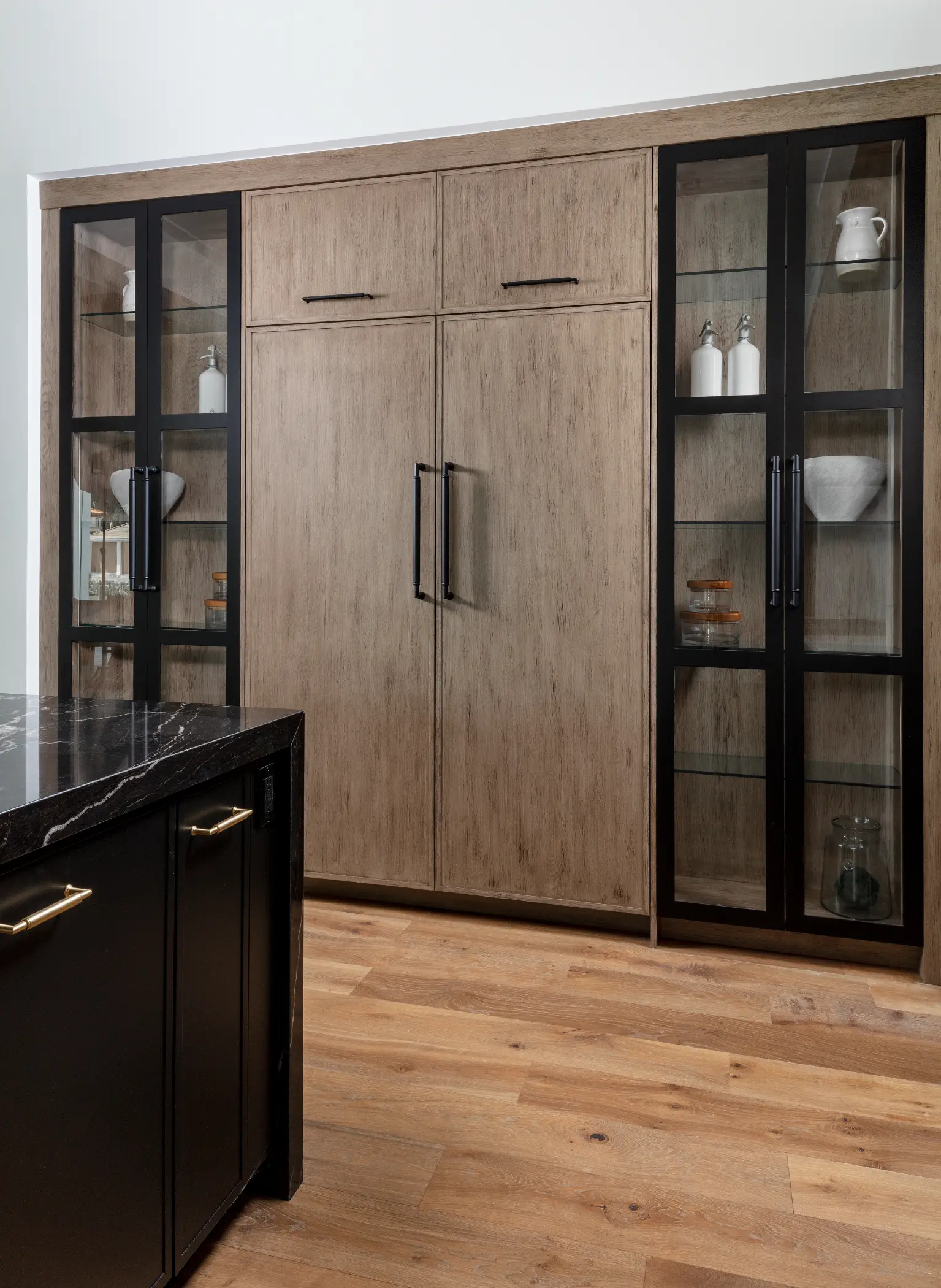 Wooden kitchen cabinets with glass doors display dishes and bottles, next to a black island.