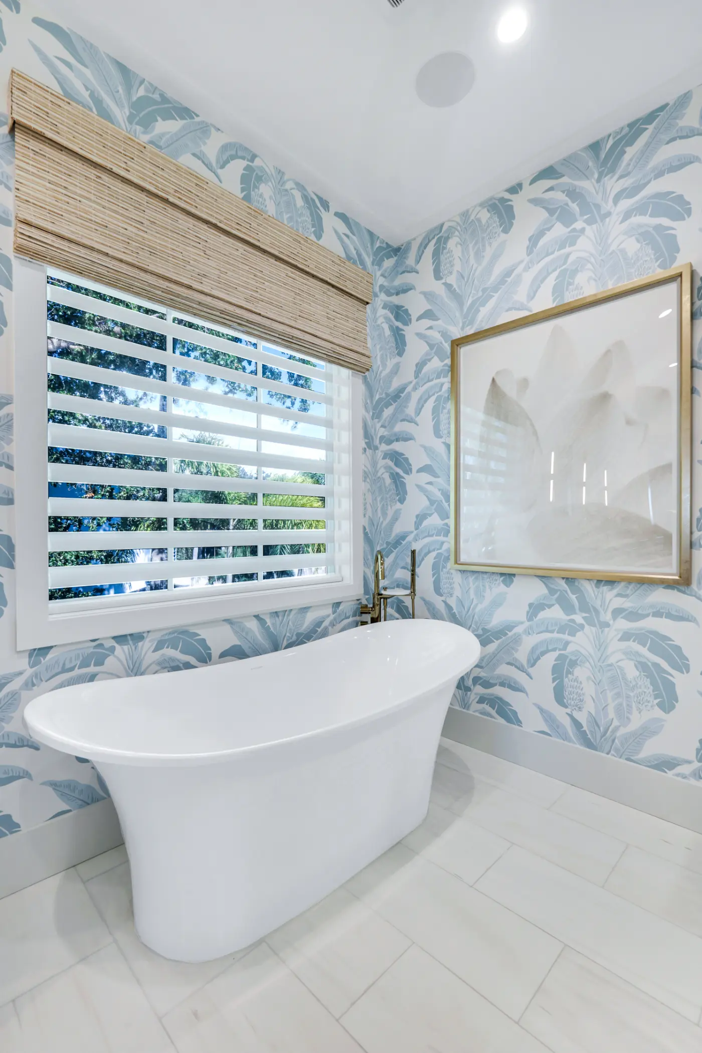 A white bathtub is placed below a window in a bathroom with floral wallpaper.