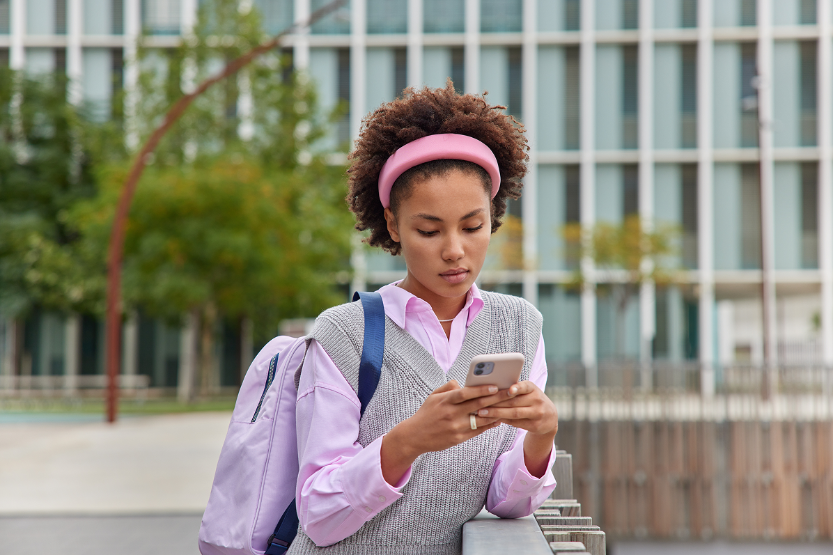 Young woman with curly hair wearing a pink headband, pink shirt, and gray sweater vest, looking at her smartphone outdoors near a railing with a lavender backpack over one shoulder and a building with vertical windows in the background.