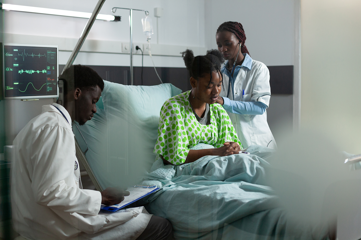 Healthcare professionals attending to a patient in a hospital room; one doctor listens to the patient's chest with a stethoscope while another records notes on a clipboard near a hospital bed with medical monitors displaying vital signs.