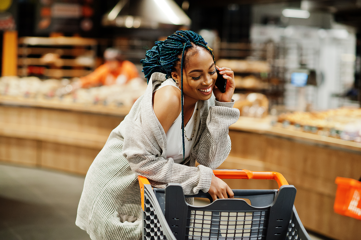 Smiling woman with blue braided hair leaning on a shopping cart and talking on her phone in a grocery store with shelves of food in the background.