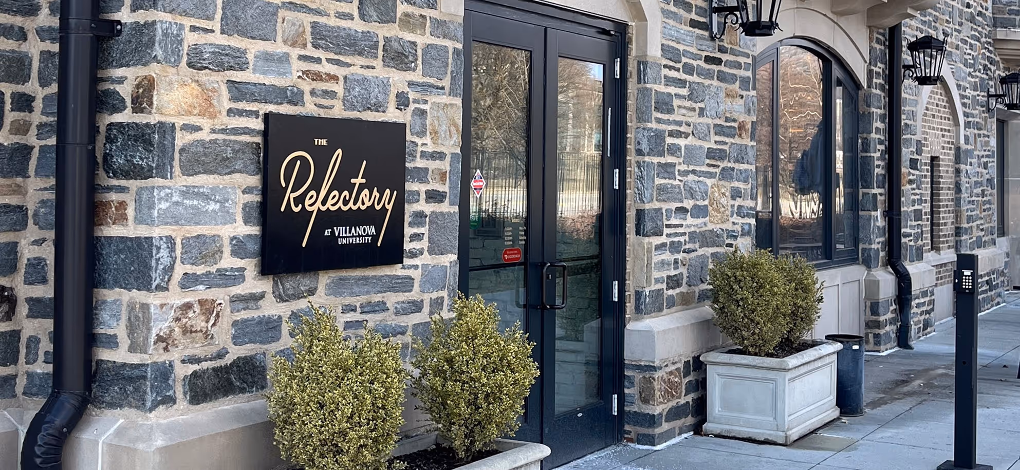 Custom outdoor business sign for The Refectory at Villanova University mounted on stone wall, featuring gold lettering on black panel