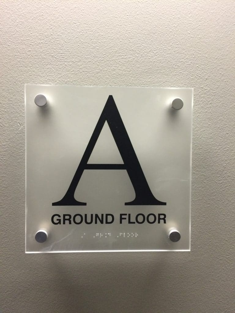 Modern ADA compliant ground floor sign with large letter A, tactile text, and braille on frosted acrylic with standoff mounts