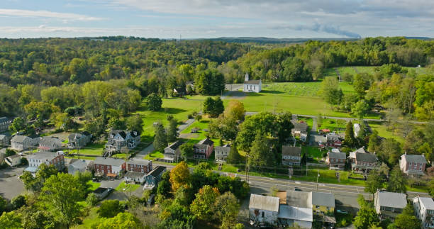 Aerial view of a small town with residential houses, a few buildings, a white church, and green forested hills in the background under a cloudy sky.