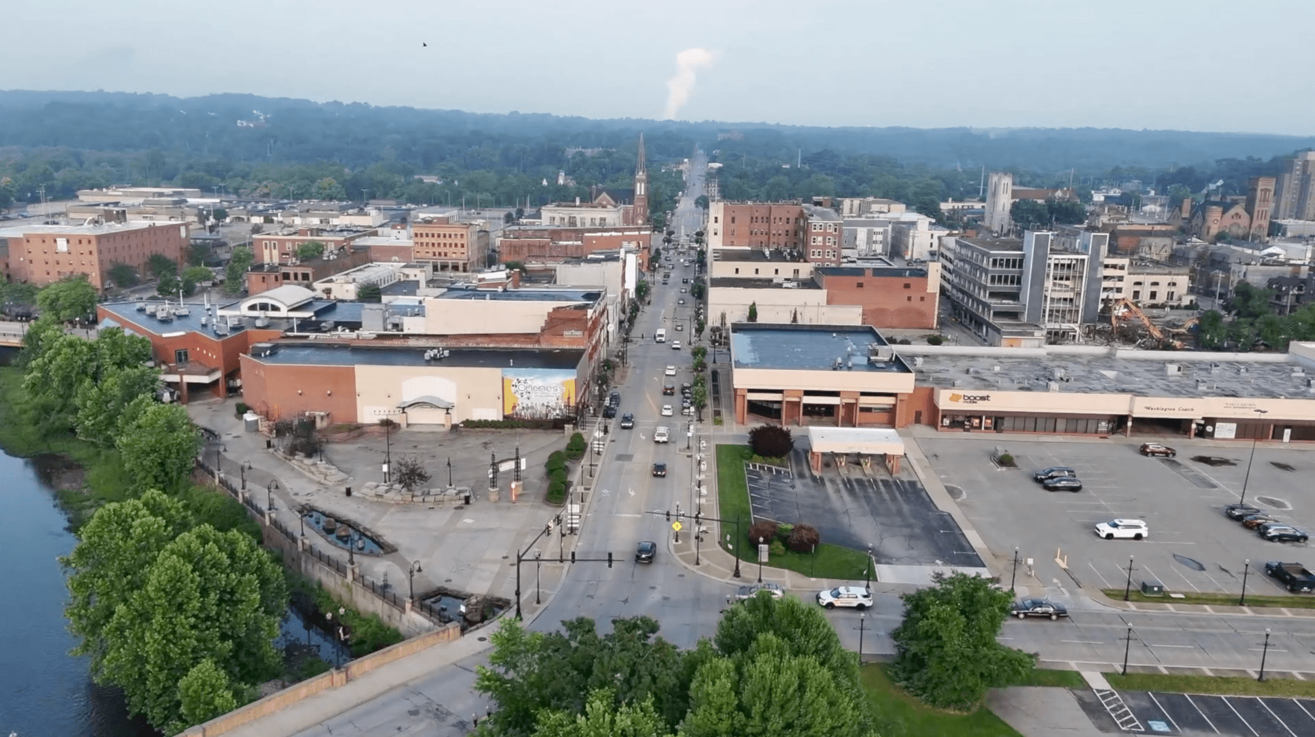 Aerial view of a mid-sized town with a main road running through commercial buildings, parking lots, and green trees alongside a river.