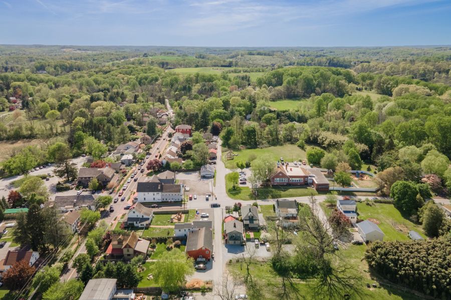 Aerial view of a small town with houses, roads, and abundant green trees extending into the rolling hills under a blue sky.