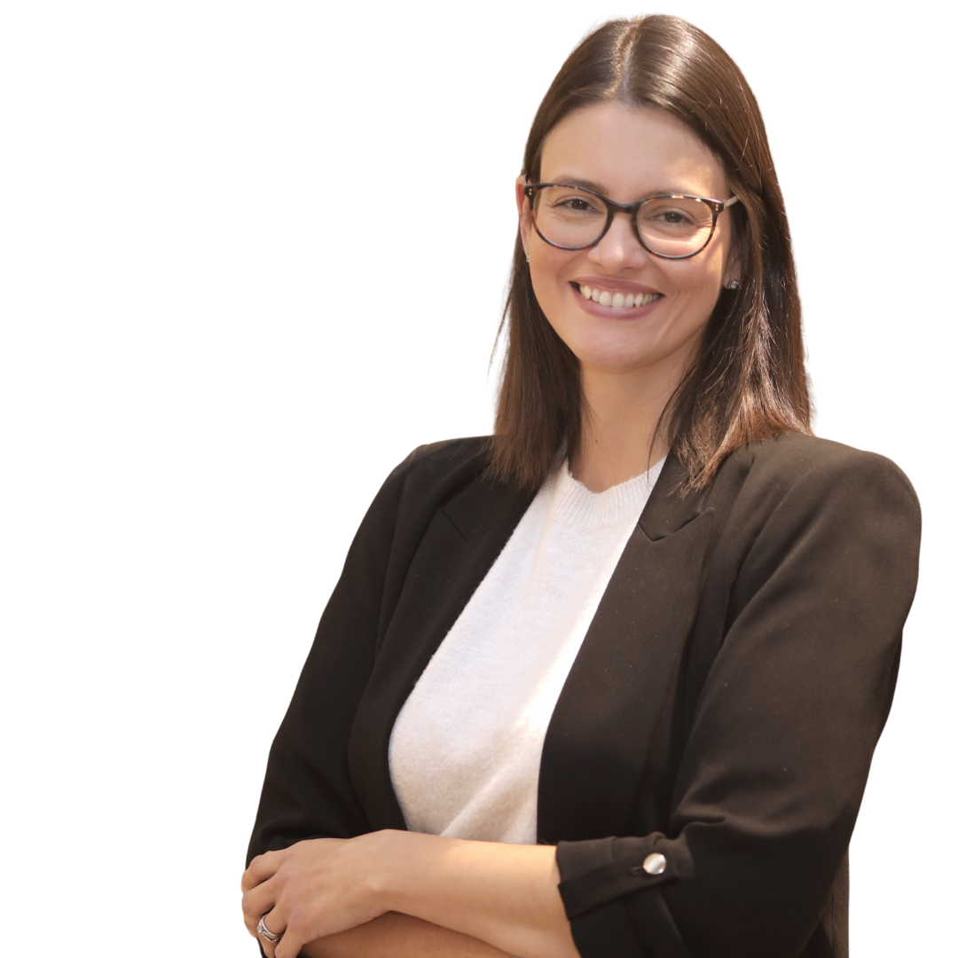 Najara, mentoria de carreira, branca, cabelos pretos, óculos de grau e sorriso no rosto. Usa blazer preto e blusa branca. Ao fundo uma estante com itens decorativos.